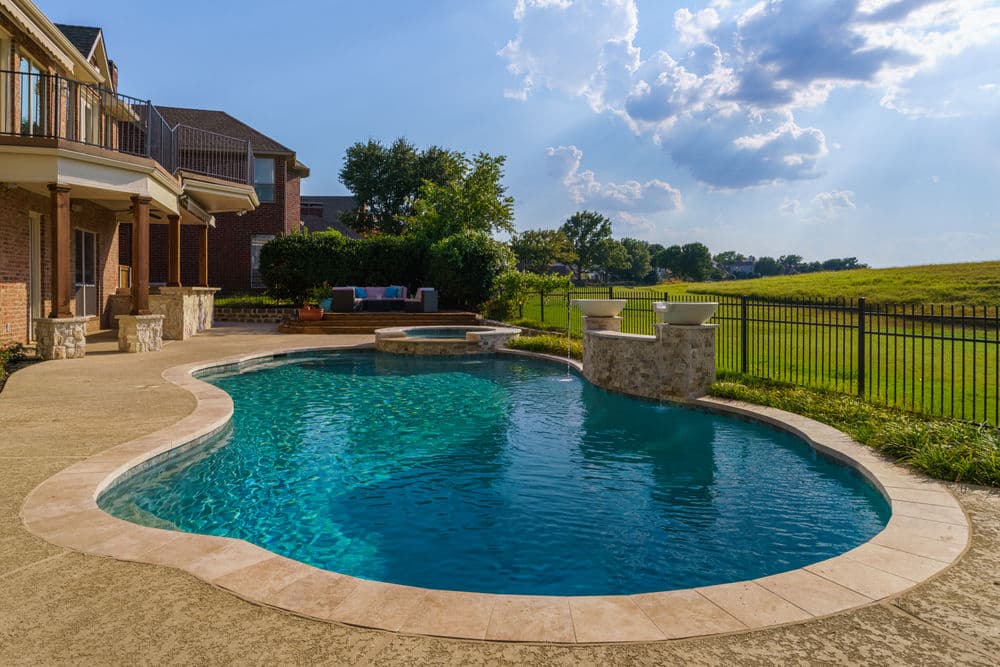 Luxury backyard pool with patio, surrounded by greenery and a clear blue sky.